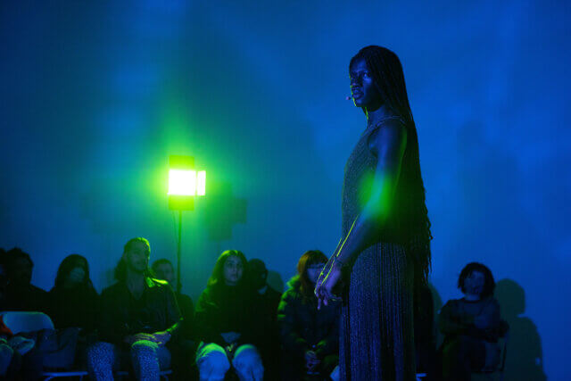 A woman with long braids and in a sequined dress stands in front of an audience. She is lit in a dark blue with a green spotlight on her.