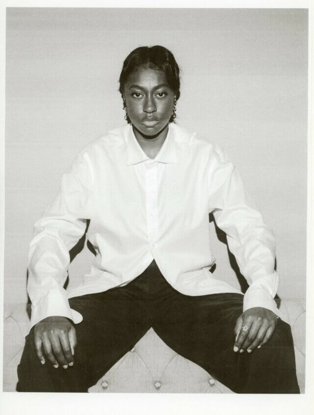 A black and white photo of a black woman in a white shirt and black trousers sitting on the back of a Chesterfield sofa.